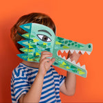 Child holding a Londji green paper crocodile mask against an orange background