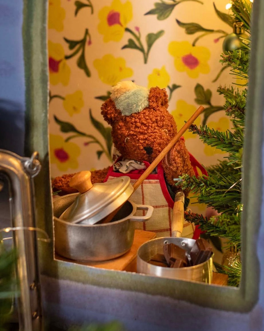Small bear figurine with a pot and spoon in front of a floral-patterned wall.