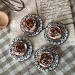 Small cinnamon rolls on black and white patterned plates with a whisk and wooden bowl on a checkered tablecloth.