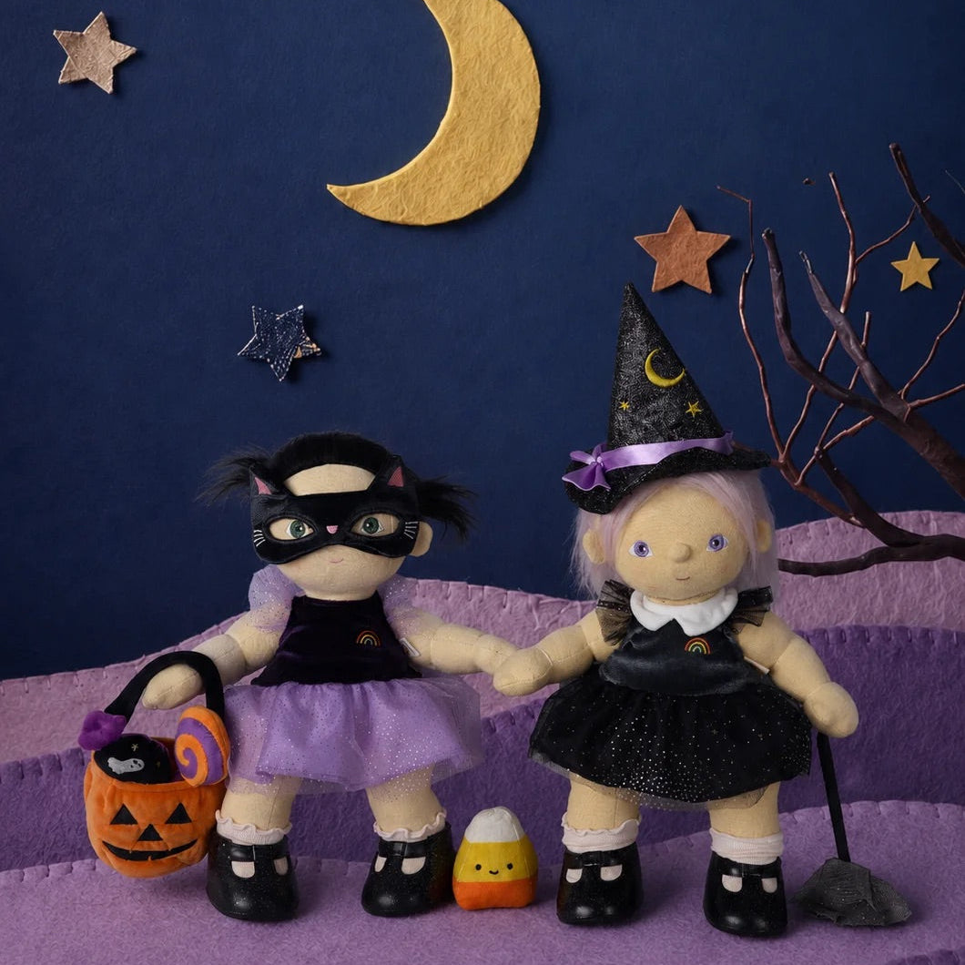 Two Halloween-themed dolls posed on a purple textured surface against a dark blue backdrop with a gold crescent moon, stars, and a bare tree branch. One doll wears a black costume with a purple skirt and holds an orange jack-o'-lantern bucket.