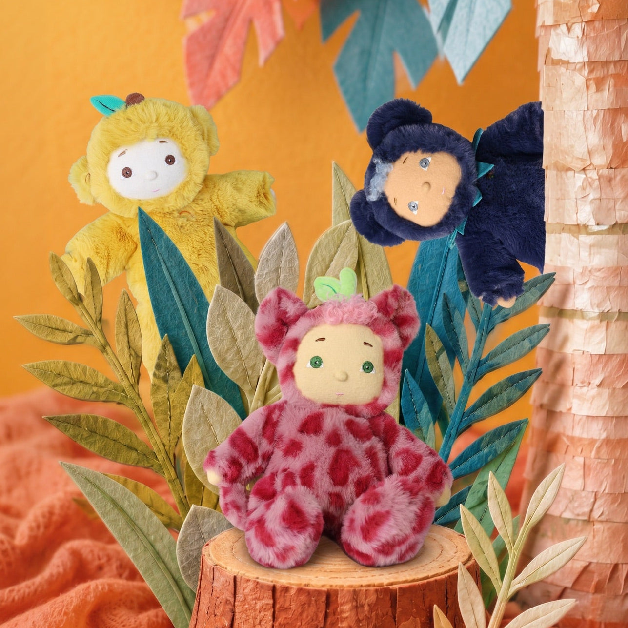 Three plush monkey toys in different colored outfits are arranged on a decorative tropical set with a faux tree trunk, paper leaves, and an orange backdrop.