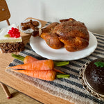 Miniature roasted chicken on a white plate with carrots and other food items on a wooden table.