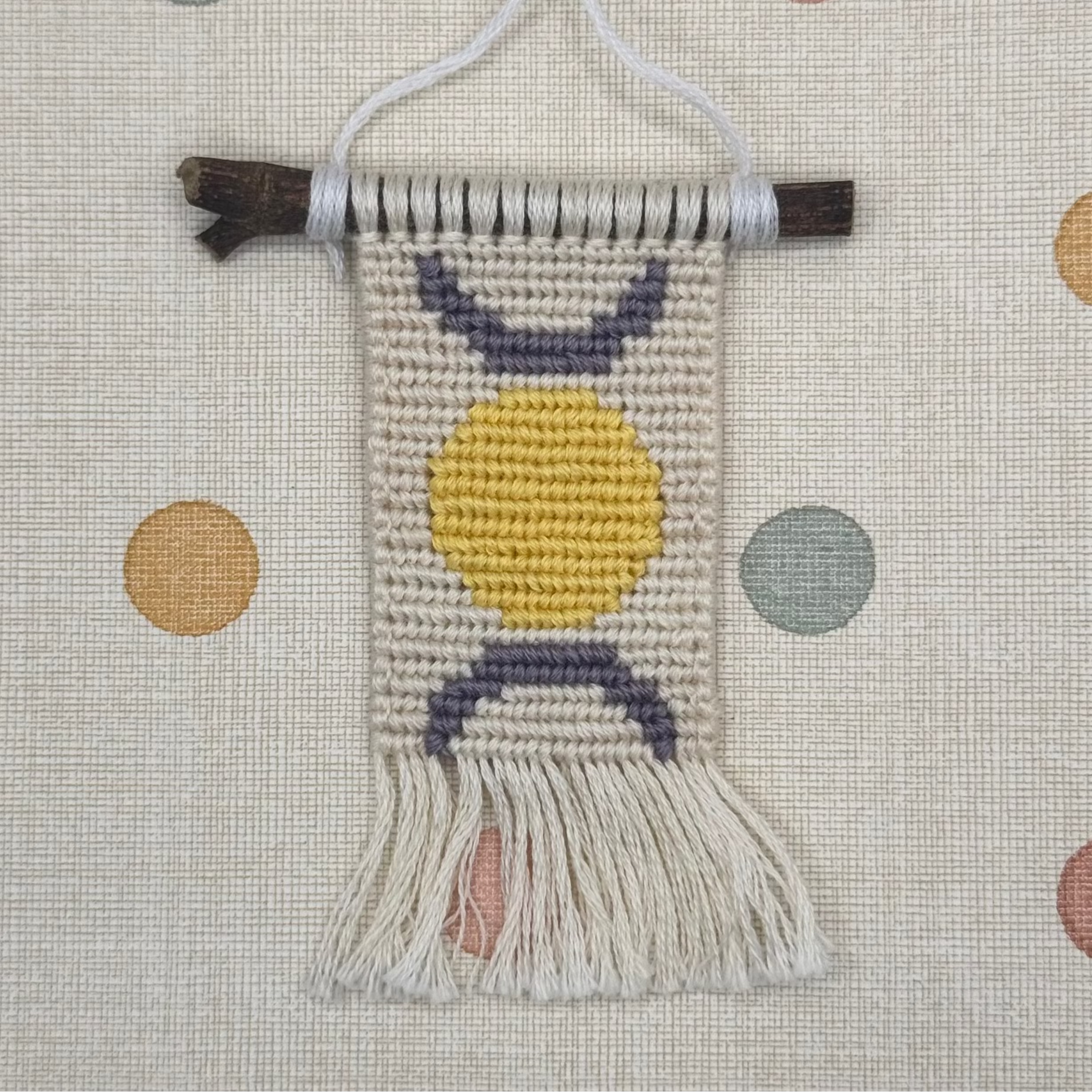 Small woven wall hanging with a yellow circle and purple arch on a polka dot background