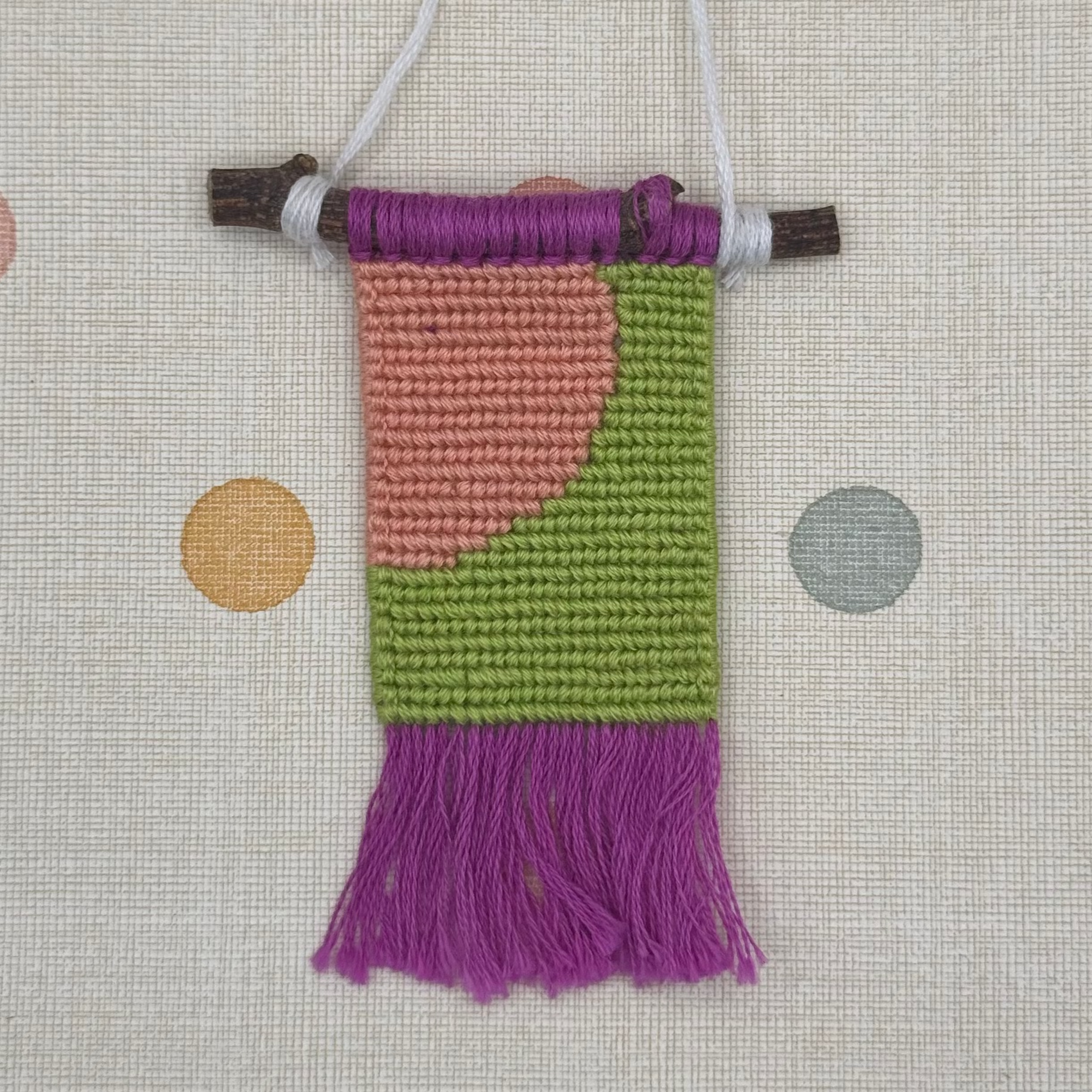 Small woven wall hanging with purple, pink, and green pattern on a polka dot background