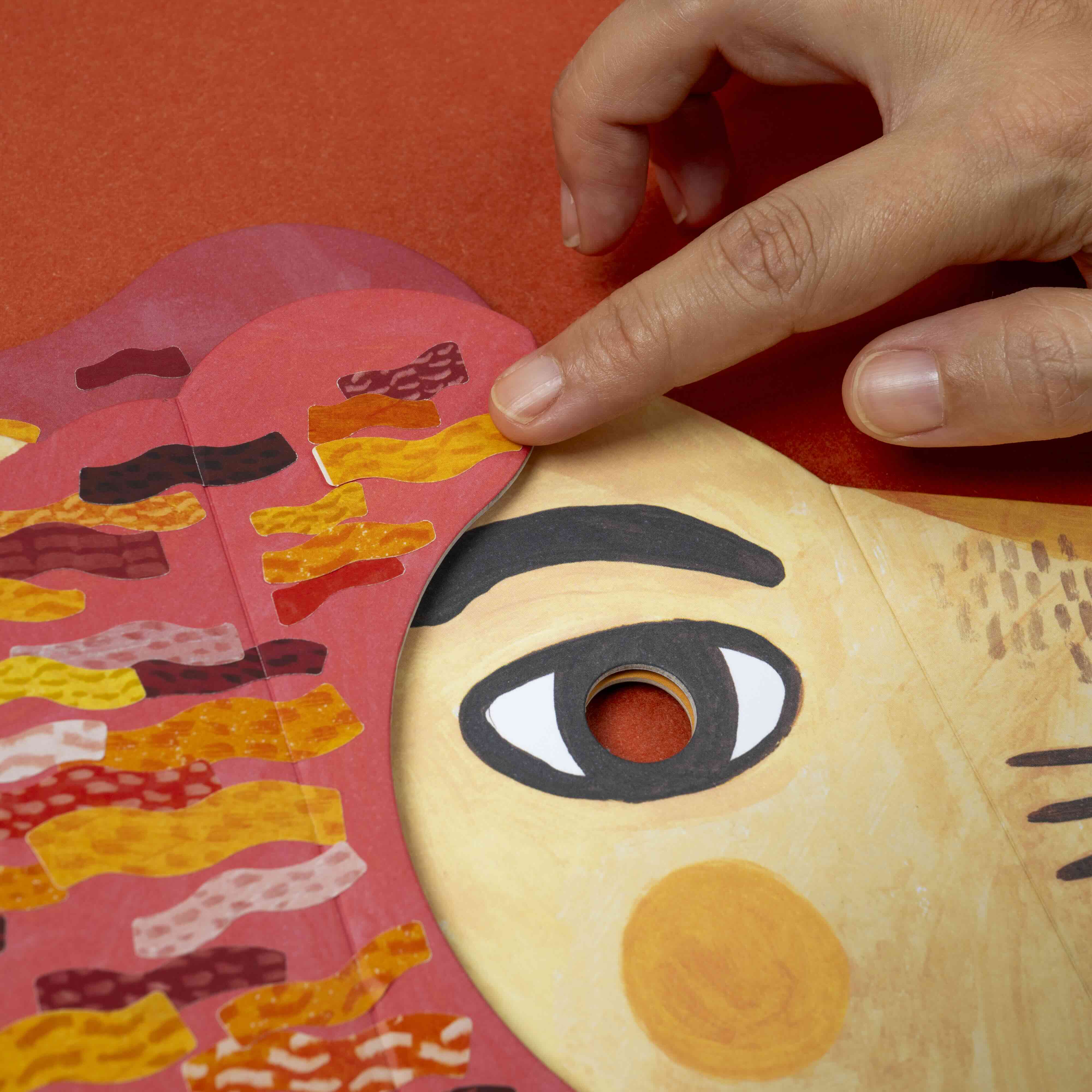 Hand interacting with a colorful paper lion mask by Londji on an orange background