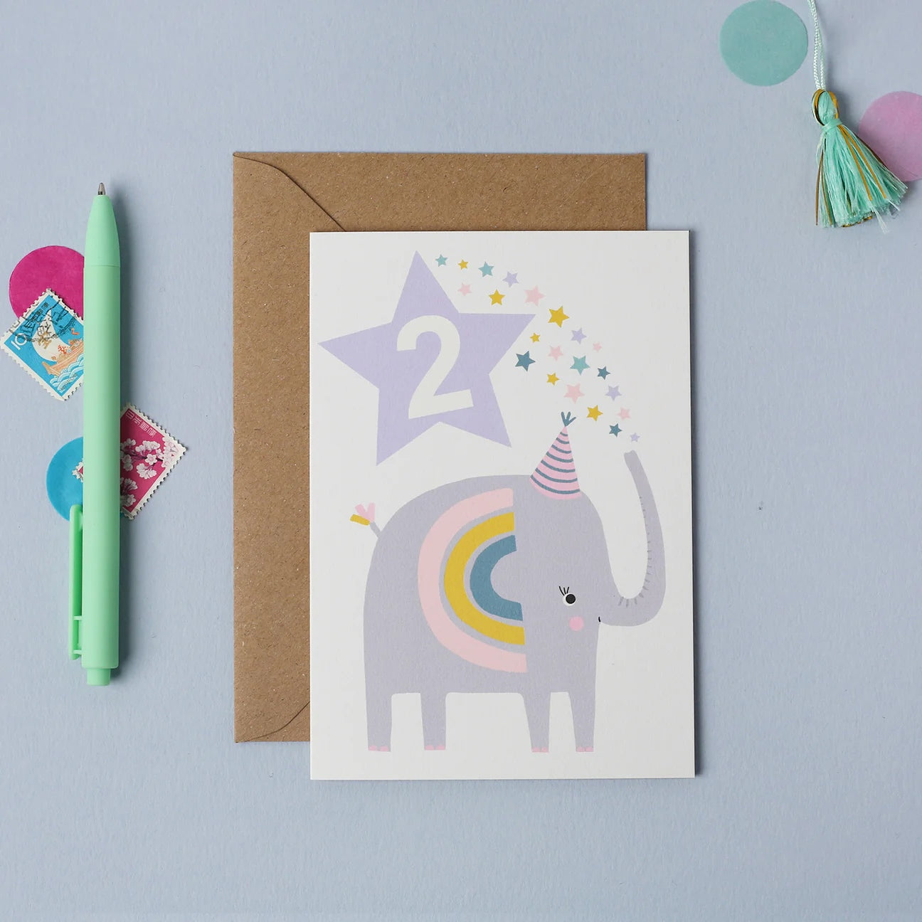 Mifkins children's birthday card with an elephant, star, and rainbow on a light blue background with a pen and envelope.