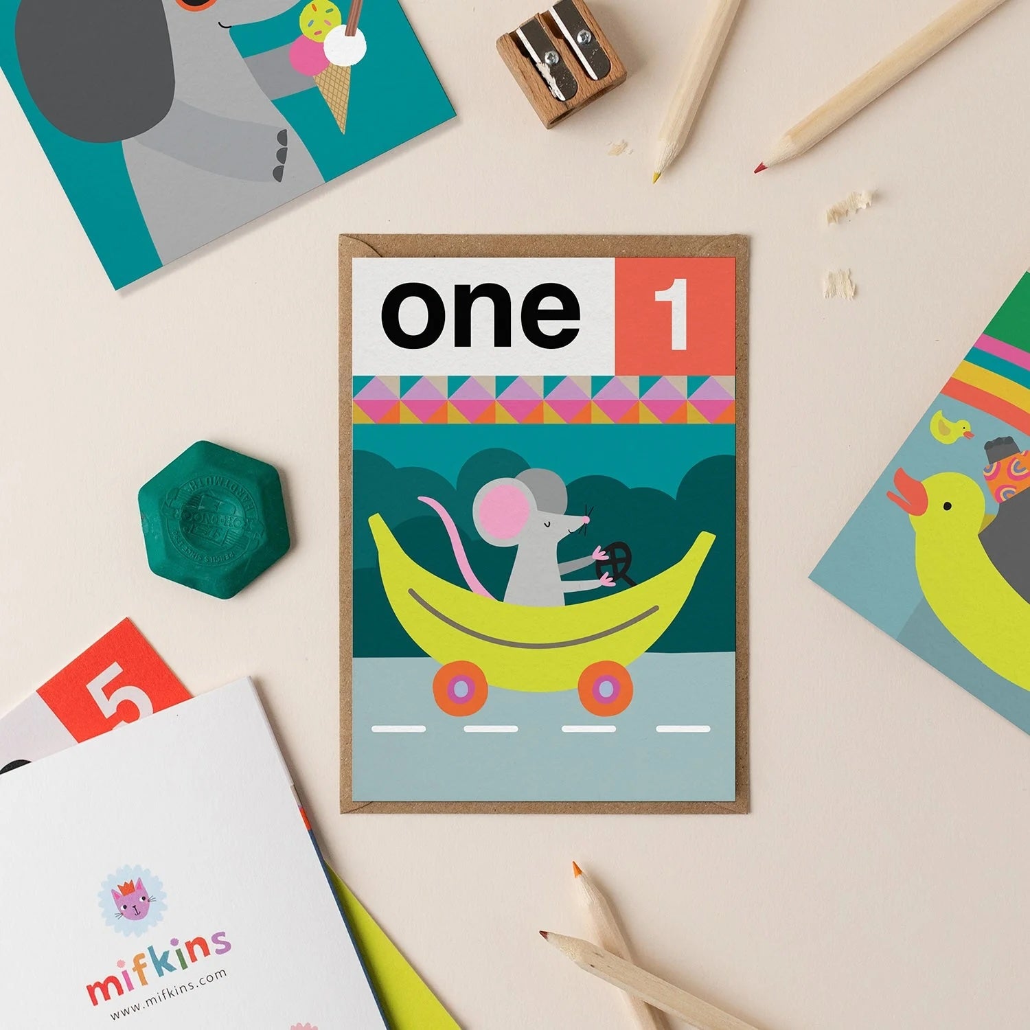 Mifkins children's card with a mouse on a banana, surrounded by stationery items on a light background.