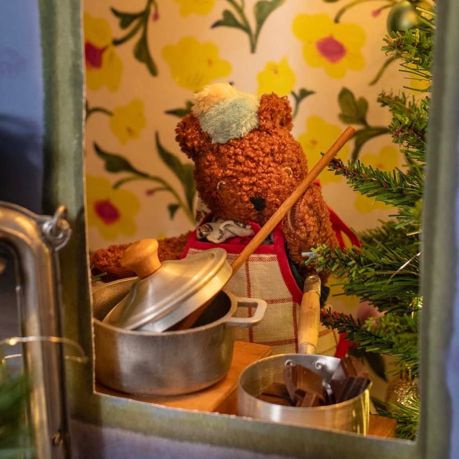 Small bear figurine with a pot and spoon in front of a floral-patterned wall.