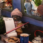 Toy cat in a festive setting with cookies, hot chocolate, and a letter, surrounded by Christmas decorations.