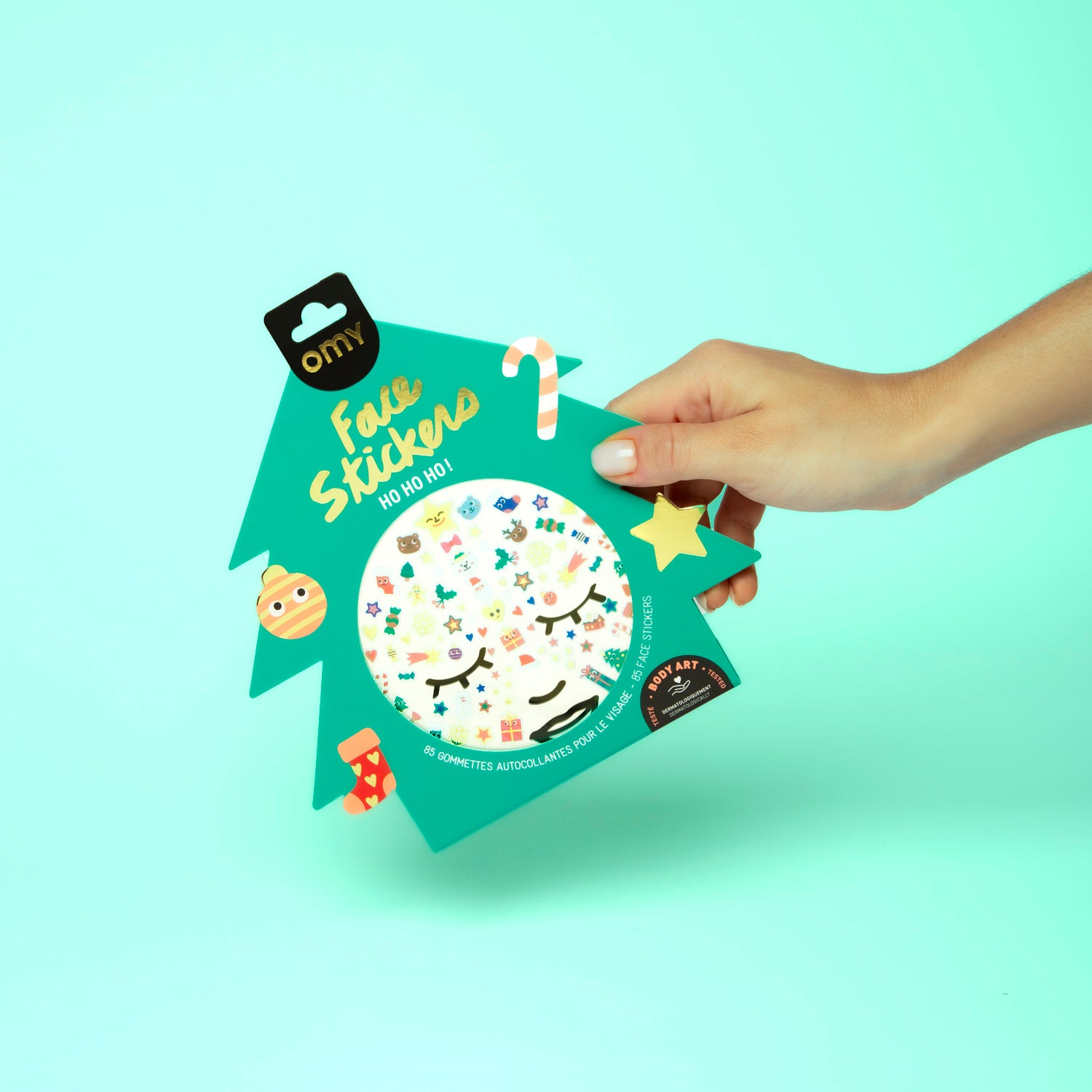 Hand holding a package of Omy face stickers against a light green background