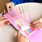 Children's hands opening a pink OMY nail sticker book with colorful stickers.