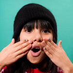 Child with black knit hat covering mouth with hands with OMY nail stickers on teal background