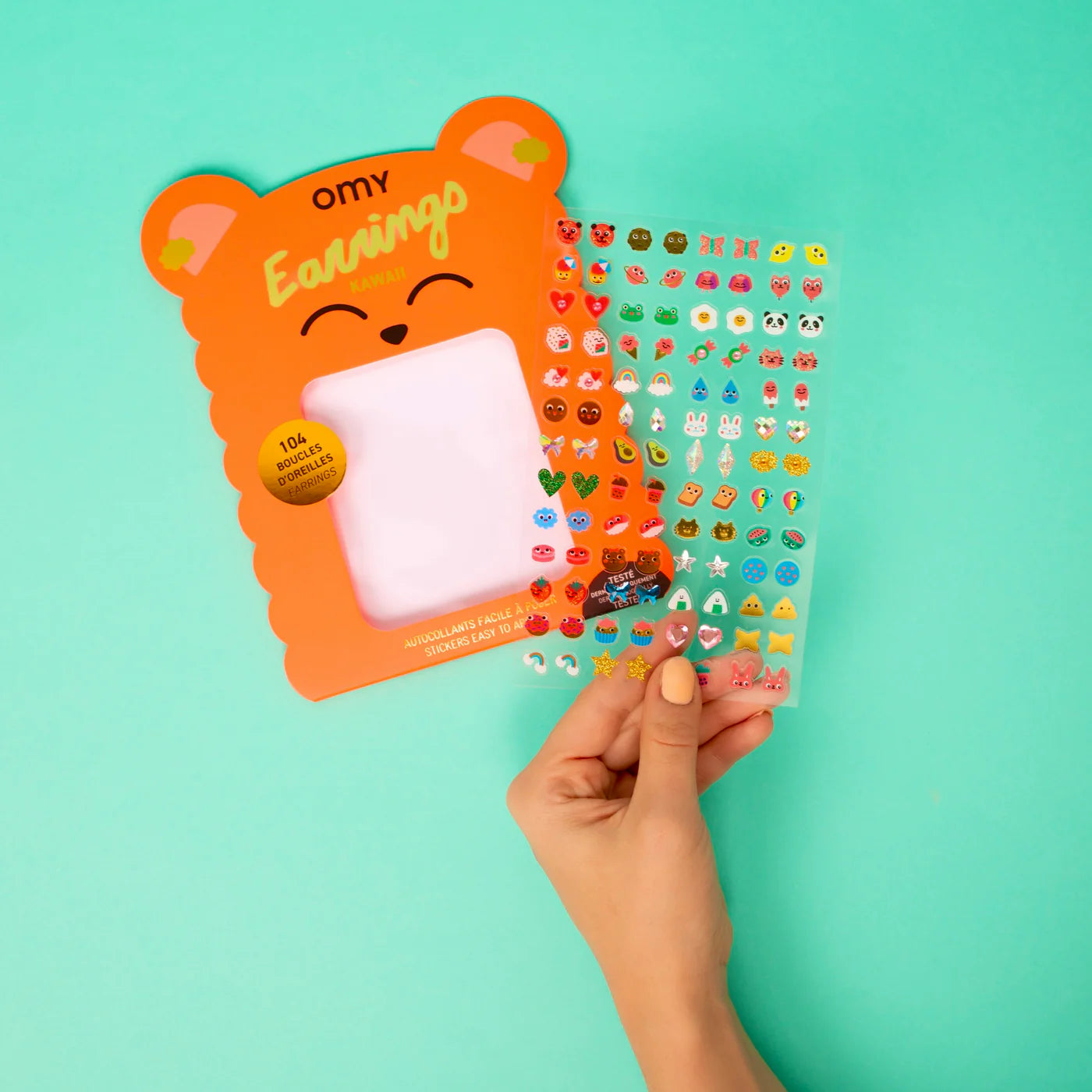 Hand holding a card with OMY bear design and colorful earrings against a teal background