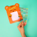 Hand holding a card with OMY bear design and colorful earrings against a teal background