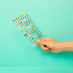 Hand holding a sheet of colorful OMY nail stickers against a turquoise background