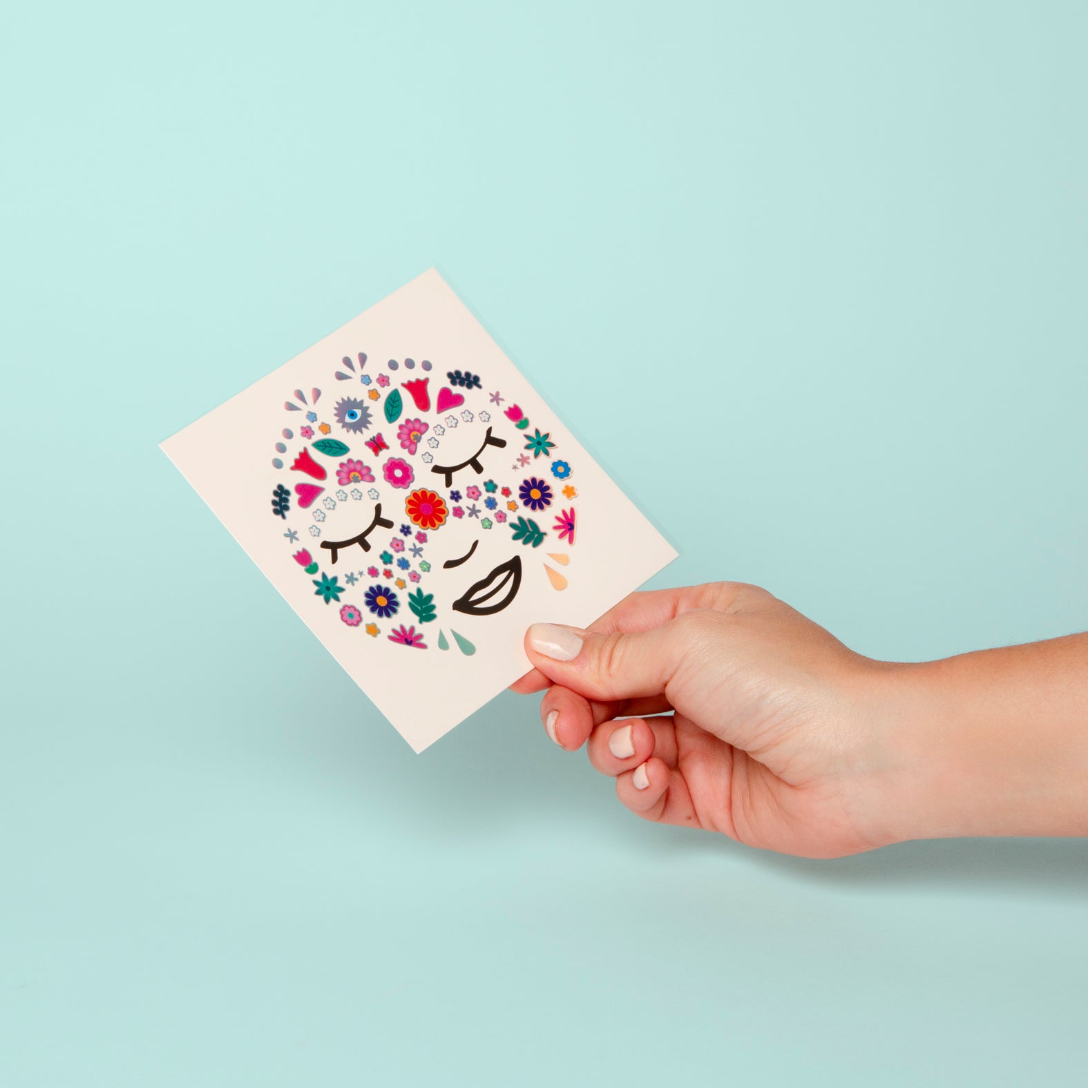Hand holding a card with OMY floral and face design against a light blue background