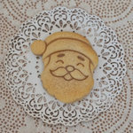 A small round golden-brown cookie shaped like Santa's face sits on a white lace doily over a beige patterned fabric background.