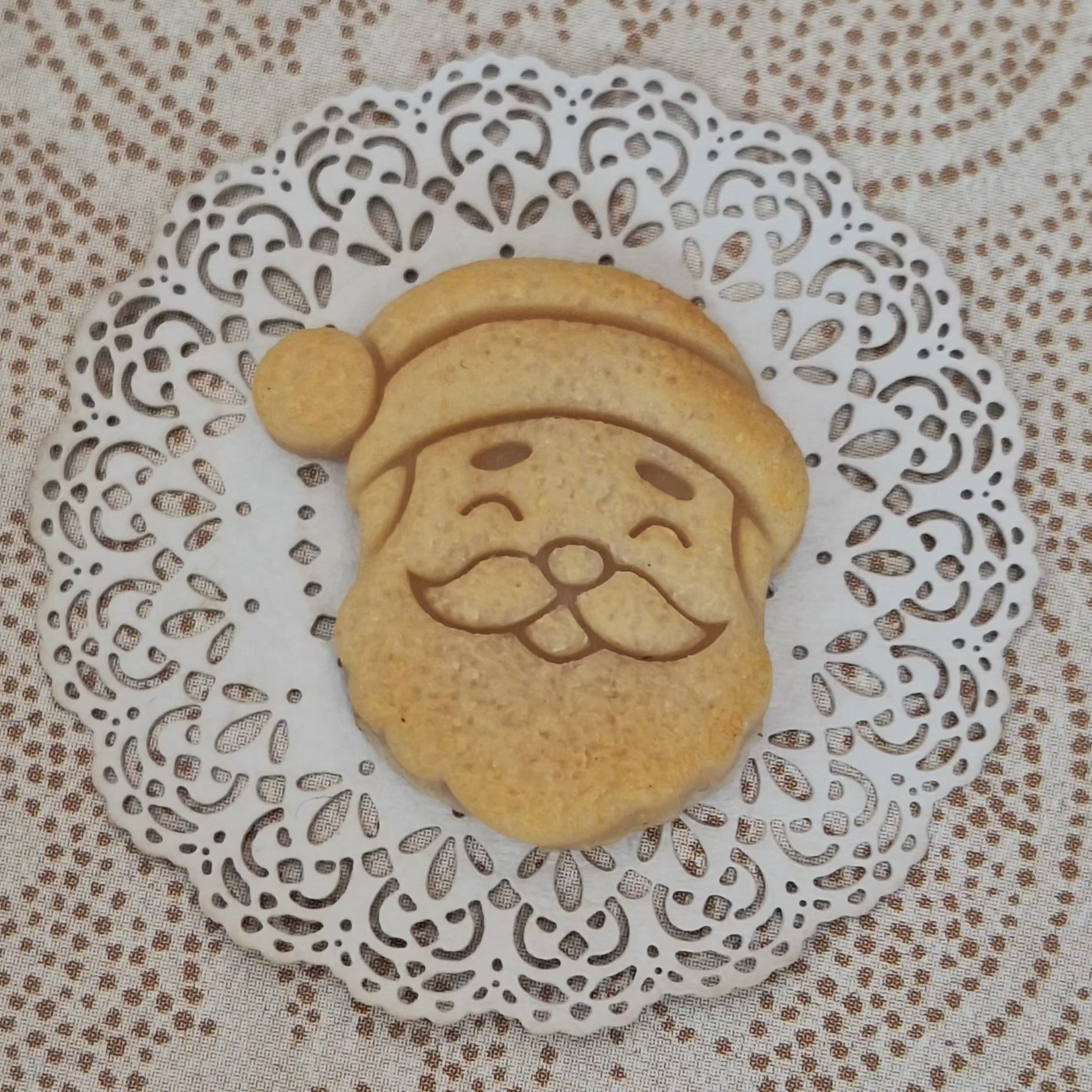 A small round golden-brown cookie shaped like Santa's face sits on a white lace doily over a beige patterned fabric background.