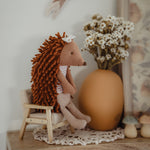 A small plush hedgehog doll with orange-brown textured spines and a white bow sits on a miniature wooden chair beside a large terracotta vase filled with white dried flowers, displayed on a wooden shelf.